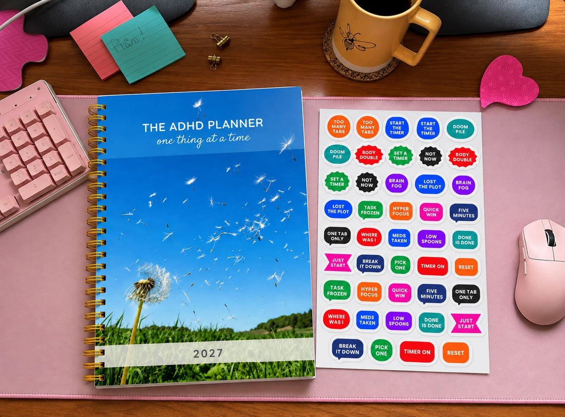 The ADHD Planner with a dandelion cover on a pink desk mat, next to a sheet of word stickers reading Too Many Tabs, Doom Pile, Body Double, Task Frozen, Just Start and Break It Down